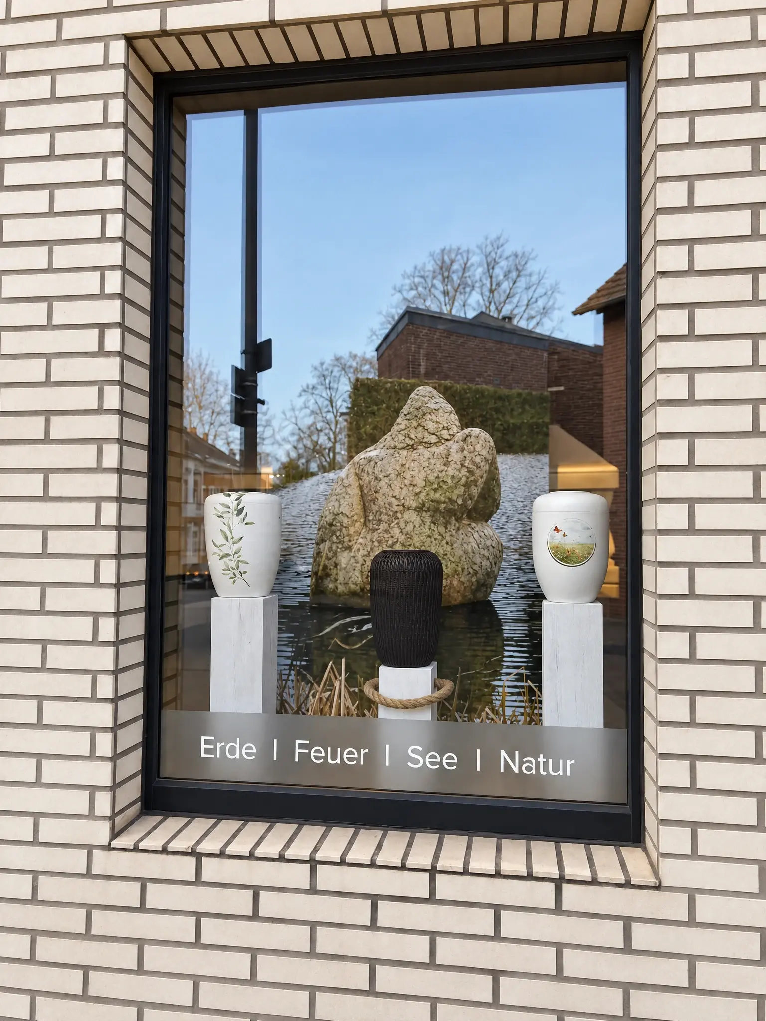 Schaufenster mit drei Urnen vor einer Steinskulptur und dem Schriftzug Erde, Feuer, See, Natur