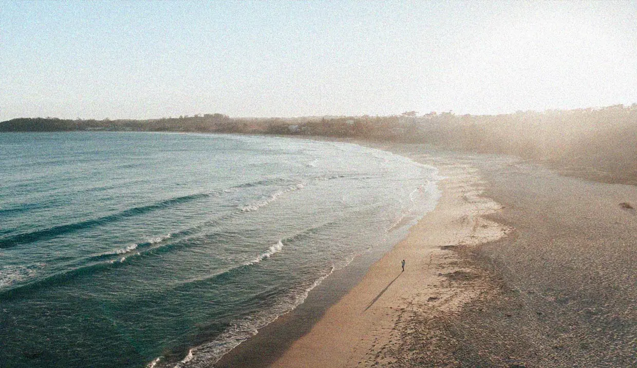 Weiter Sandstrand im Abendlicht, an dem ein einzelner Mensch am Wasser entlanggeht